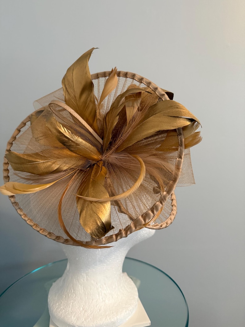May include: A gold-colored fascinator with a circular base and layered design. The headpiece features large, stylized feathers and a sheer, net-like material. The hat is displayed on a white mannequin head.