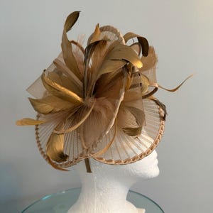 May include: A tan and gold fascinator with a circular base and a headband. The headpiece features layers of tan netting and gold-colored feathers arranged in a radial pattern. The base is a circular design with a decorative edge.