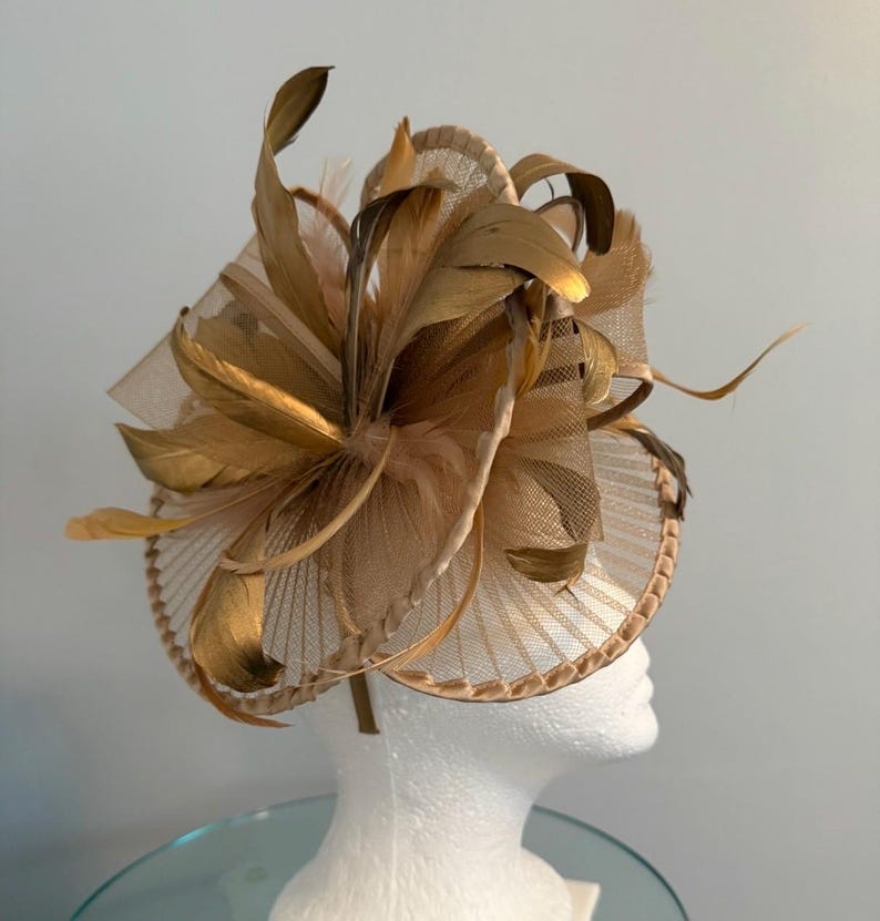 May include: A tan and gold fascinator with a circular base and a headband. The headpiece features layers of tan netting and gold-colored feathers arranged in a radial pattern. The base is a circular design with a decorative edge.