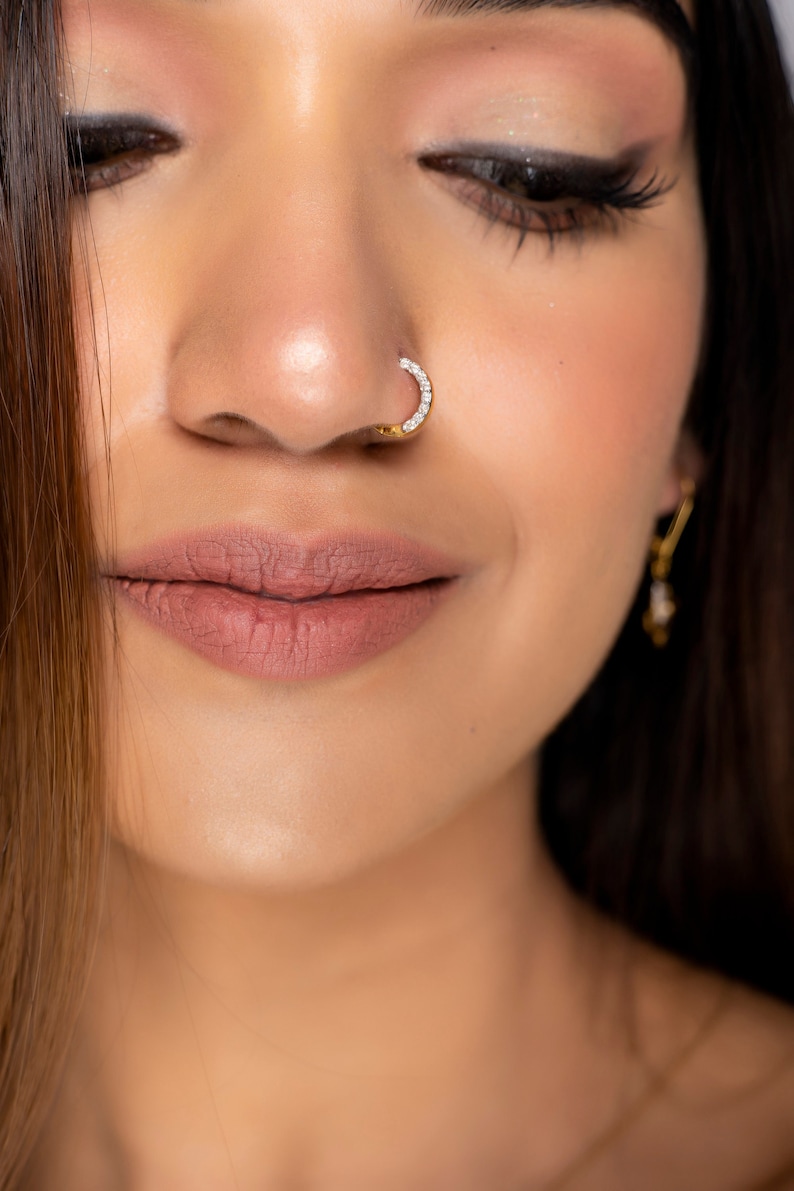 May include: Close-up of a woman's face with a gold nose ring set with small clear stones. The ring is a simple, curved design.
