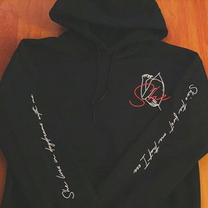 Könnte beinhalten: Schwarzer Hoodie mit einem roten und weißen Grafikdruck auf der Vorderseite und weißem Text auf den Ärmeln. Der Text auf dem linken Ärmel lautet "She lives in defiance with me" und der Text auf dem rechten Ärmel lautet "Since the first one I have lost my soul".