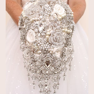 Peut inclure: Un bouquet de mariée composé de roses blanches en satin et de perles, rehaussé d'une grande broche en strass en cascade.