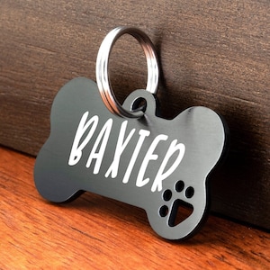 May include: Black bone-shaped dog tag with a silver ring and the name "Baxter" engraved in white lettering. A paw print is also engraved on the tag.