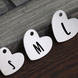 May include: Three silver heart-shaped tags with the letters S, M, and L engraved on them. The tags have a hole at the top for hanging.