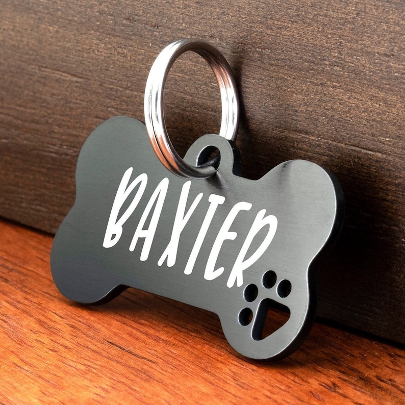 May include: Black bone-shaped dog tag with a silver ring and the name "Baxter" engraved in white lettering. A paw print is also engraved on the tag.