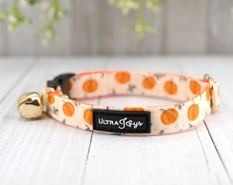 May include: A white and orange cat collar with a pumpkin print and a gold bell. The collar has a black buckle and a black label that says "Ultra Joys".