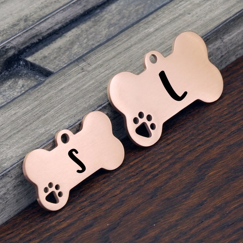 May include: Two rose gold bone-shaped dog tags with paw print designs. The tag on the left has the letter "S" engraved on it. The tag on the right has the letter "L" engraved on it.