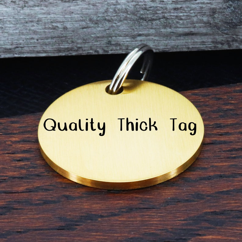 May include: A round, gold-colored metal dog tag with a silver key ring. The tag is engraved with the words "Quality Thick Tag".