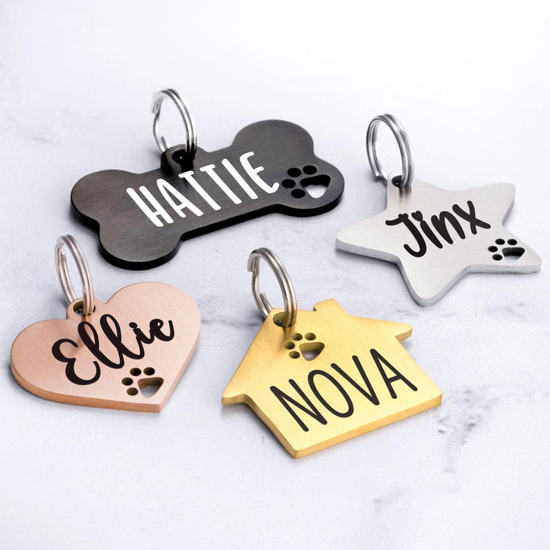 May include: Four personalized dog tags in different shapes and colors. A black bone tag with the name "Hattie", a silver star tag with the name "Jinx", a rose gold heart tag with the name "Ellie", and a gold house tag with the name "Nova".