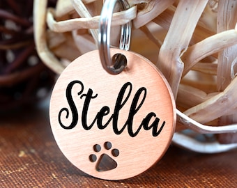 May include: Copper dog tag with the name "Stella" engraved in black cursive font. The tag has a paw print cutout and a silver key ring.