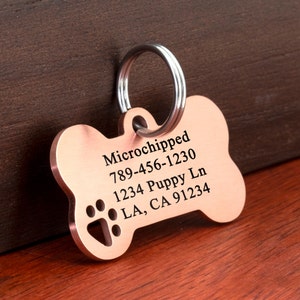 May include: A copper bone-shaped dog tag with a silver ring. The tag is engraved with the text "Microchipped 789-456-1230 1234 Puppy Ln LA, CA 91234" and a paw print.