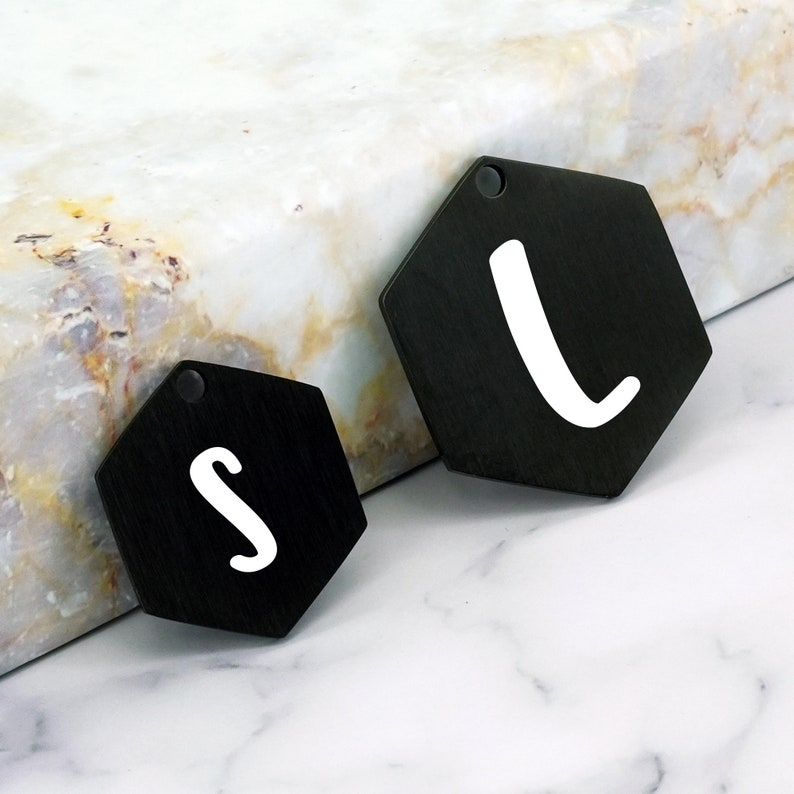 May include: Two black metal hexagon-shaped charms with white lettering. The larger charm has the letter "L" and the smaller charm has the letter "S".