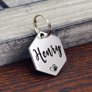 May include: Silver hexagon-shaped dog tag with a silver ring. The tag is engraved with the name "Henry" and a heart with a paw print.