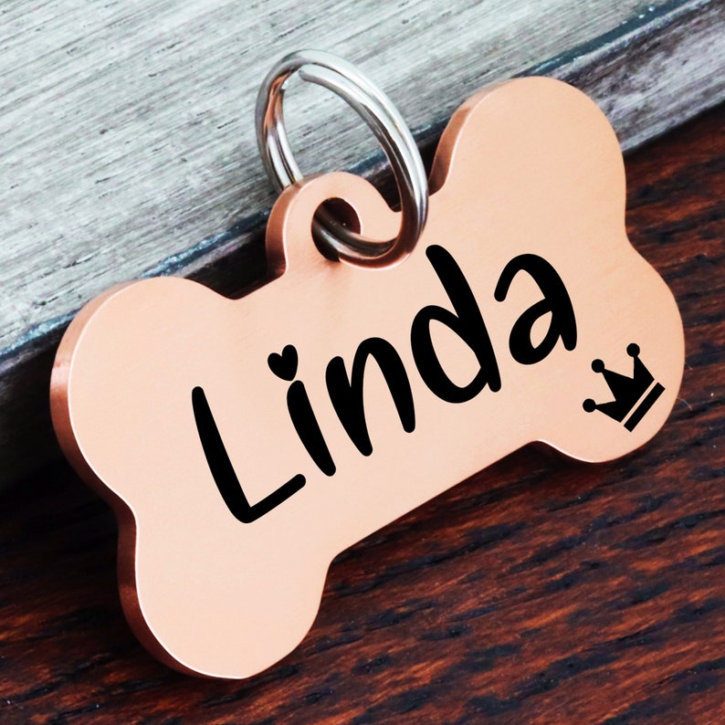 May include: A rose gold bone-shaped dog tag with the name "Linda" engraved in black lettering and a small crown symbol.