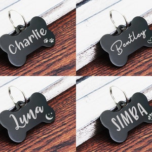 May include: Four black bone-shaped dog tags with engraved names and paw prints. The tags are on a wooden surface. The names on the tags are Charlie, Bentley, Luna, and Simba.