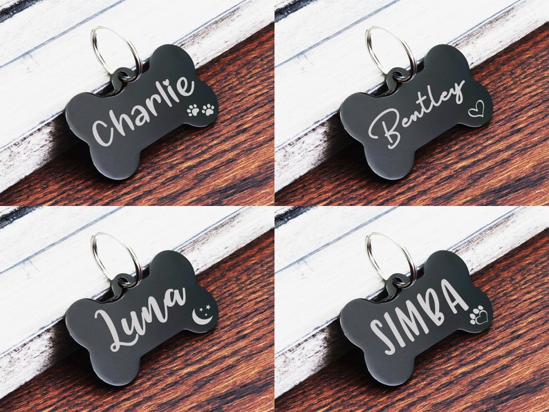 May include: Four black bone-shaped dog tags with engraved names and paw prints. The tags are on a wooden surface. The names on the tags are Charlie, Bentley, Luna, and Simba.