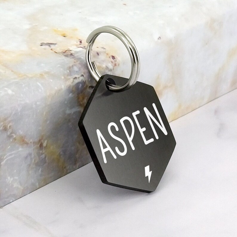 May include: Black hexagonal pet ID tag with a silver ring and the name "ASPEN" in white lettering with a lightning bolt symbol.
