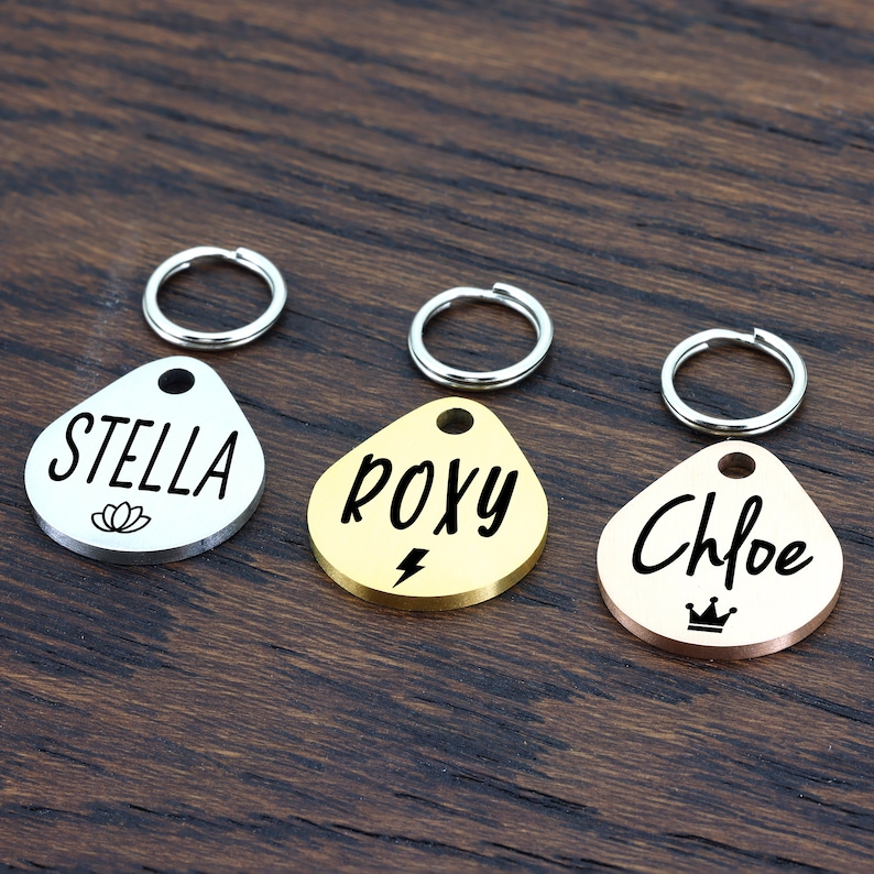 May include: Three personalized pet ID tags in the shape of a teardrop. The tags are silver, gold, and rose gold. The silver tag says "STELLA" with a flower design, the gold tag says "ROXY" with a lightning bolt design, and the rose gold tag says "Chloe" with a crown design.