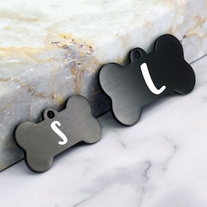 May include: Two black bone-shaped dog tags with white letters. The tag on the left says "S" and the tag on the right says "L".
