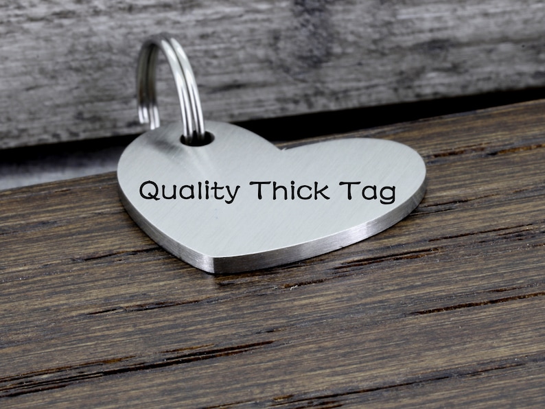 May include: A silver heart-shaped tag with a keyring attached. The tag is engraved with the text "Quality Thick Tag".