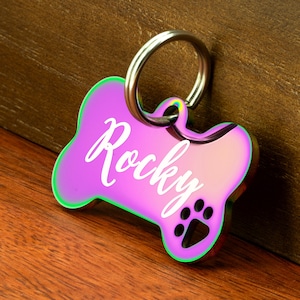 May include: A pink iridescent dog tag with a bone shape and the name "Rocky" engraved in white. The tag has a silver metal ring for attaching to a collar.
