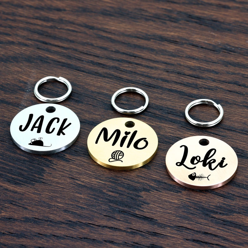 May include: Three round pet ID tags with different colored metal finishes. The tags are engraved with the names "Jack", "Milo", and "Loki" and each has a small icon. The Jack tag is silver with a mouse icon, the Milo tag is gold with a yarn ball icon, and the Loki tag is rose gold with a fish bone icon.