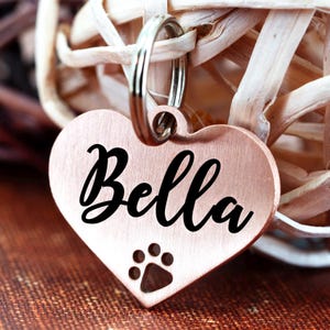 May include: A heart-shaped copper dog tag with a paw print cutout and the name "Bella" engraved in black lettering.