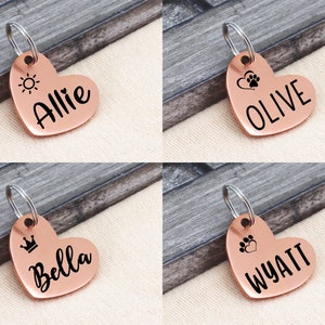 May include: Four copper heart-shaped dog tags with different engravings. The tags feature a sun, paw prints, a crown, and a heart. The names Allie, Olive, Bella, and Wyatt are engraved on the tags.