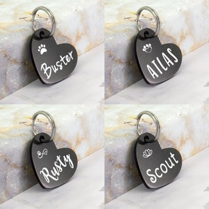 May include: Four black heart-shaped dog tags with silver rings. The tags are engraved with the names "Buster", "Atlas", "Rusty", and "Scout". Each tag also features a small design, such as a paw print, a heart, an infinity symbol, or a flower.