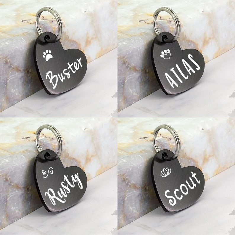 May include: Four black heart-shaped dog tags with silver rings. The tags are engraved with the names "Buster", "Atlas", "Rusty", and "Scout". Each tag also features a small design, such as a paw print, a heart, an infinity symbol, or a flower.