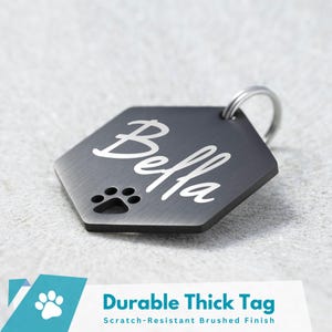 May include: A black hexagonal dog tag with a brushed finish. The tag has a paw print cutout and the name "Bella" engraved in a silver font.