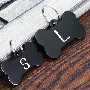 May include: Two black bone-shaped dog tags with silver rings. The tag on the left has the letter "S" engraved on it. The tag on the right has the letter "L" engraved on it.