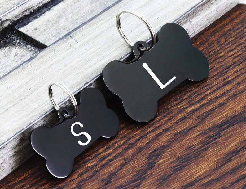 May include: Two black bone-shaped dog tags with silver rings. The tag on the left has the letter "S" engraved on it. The tag on the right has the letter "L" engraved on it.