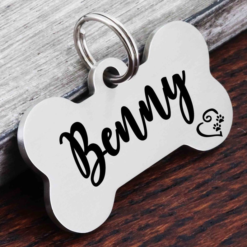 May include: Silver bone-shaped dog tag with the name "Benny" engraved in black script. A small heart with paw prints is engraved on the right side of the tag.