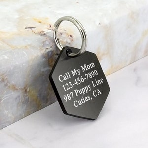 May include: Black metal dog tag with engraved text: "Call My Mom 123-456-7890 987 Puppy Line Cutie's, CA". The tag has a silver metal ring attached.
