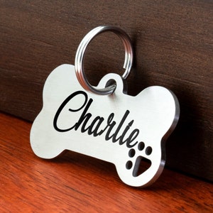May include: Silver bone-shaped dog tag with a ring for attaching to a collar. The tag has the name "Charlie" engraved on it with a paw print.