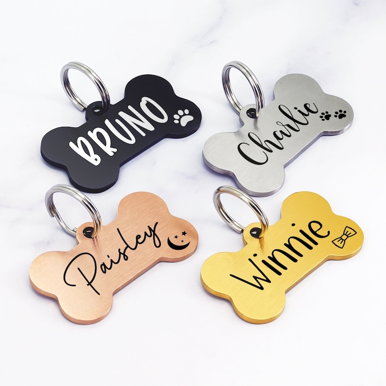 May include: Four personalized dog bone-shaped tags with different names and designs. The tags are black, silver, rose gold, and gold. The names on the tags are Bruno, Charlie, Paisley, and Winnie.