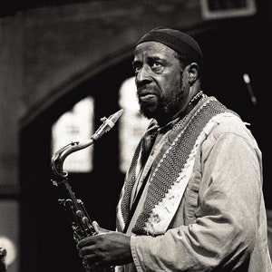 Puede incluir: Fotografía en blanco y negro de un hombre tocando el saxofón. El hombre lleva una gorra oscura, una bufanda estampada y una camisa de manga larga. El saxofón es de latón, con un cuello curvo y una campana grande.