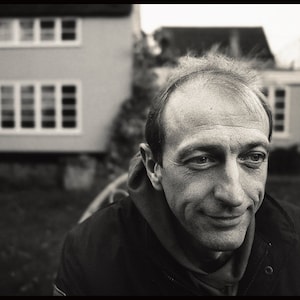 May include: Monochrome portrait of a man smiling, with a house in the background. The man is wearing a dark jacket and a hooded top. The house has several windows.
