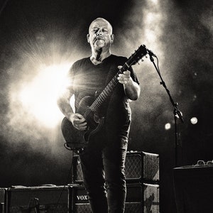Peut inclure: Image en noir et blanc d'un musicien jouant d'une guitare électrique sur scène. Le musicien porte un t-shirt noir et un pantalon sombre. L'éclairage de scène crée un halo lumineux derrière lui.