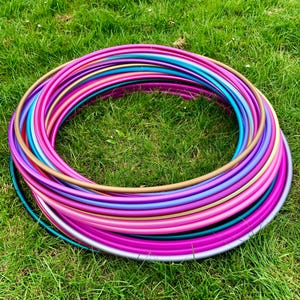 May include: A stack of colorful plastic hula hoops in various shades of pink, purple, blue, and gold. The hoops are arranged in a circle on a green grassy surface.