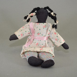 May include: A handmade rag doll with a black fabric head, arms, and legs. The doll is wearing a white dress with pink flowers and a pink ribbon. The doll has black yarn hair in pigtails.
