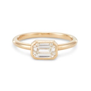 Könnte beinhalten: Ein goldener Ring mit einem Diamanten im Smaragdschliff. Der Diamant ist in einer Zargenfassung gefasst, und das Band ist aus glattem, poliertem Gold. Der Ring wird vor einem weißen Hintergrund präsentiert, um sein elegantes Design hervorzuheben. Ein klassisches Schmuckstück.