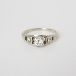 May include: A silver engagement ring with a round, clear centre stone and two smaller stones on either side.