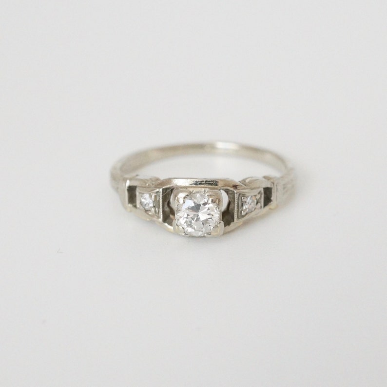 May include: A silver engagement ring with a round, clear centre stone and two smaller stones on either side.