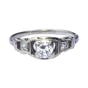 May include: A silver engagement ring featuring a round, clear centre stone and two smaller, clear stones on either side. The ring has a delicate, intricate design.
