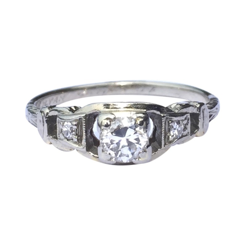 May include: A silver engagement ring featuring a round, clear centre stone and two smaller, clear stones on either side. The ring has a delicate, intricate design.