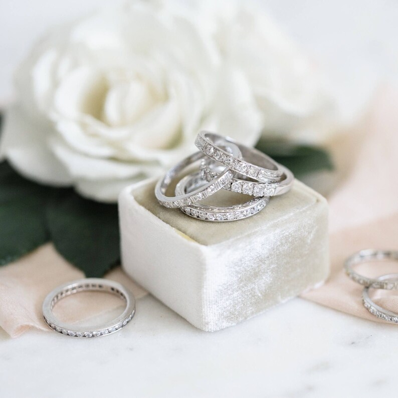 Op de afbeelding: Stapel van vier zilveren diamanten eeuwigheidsringen in een witte fluwelen ringendoos. De ringen zijn in een cirkel gerangschikt, met de grootste ring onderaan en de kleinste ring bovenaan.