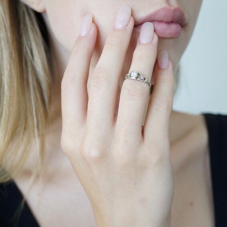May include: A close-up of a hand wearing a silver ring with a single round diamond and smaller diamonds on the band.