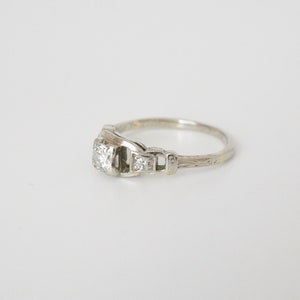 May include: A silver engagement ring featuring a single round diamond and two smaller diamonds set on the band. The band is engraved with a delicate pattern.